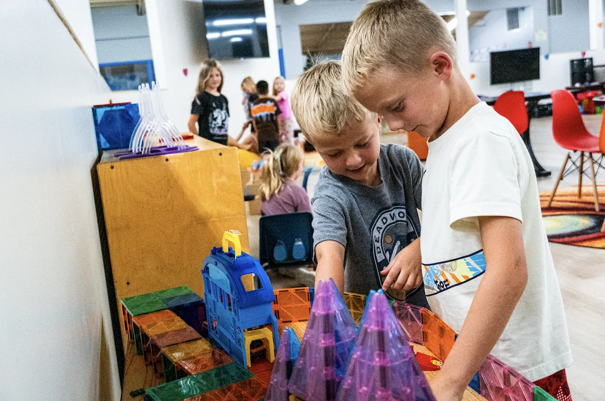 Two boys building together with magnetic tiles while other kids play nearby