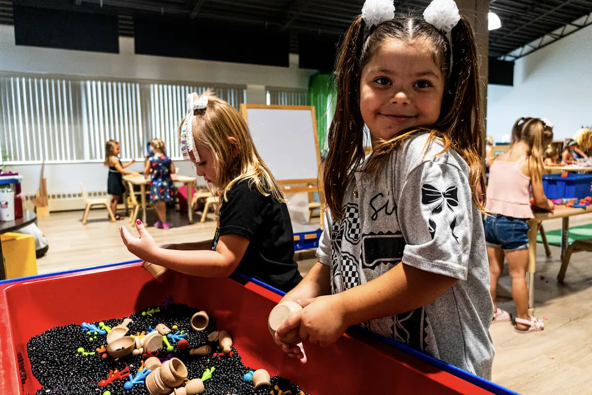 Two preschool girls explore a red sensory bin filled with wooden bowls and colorful toys at a Learning Korner activity table