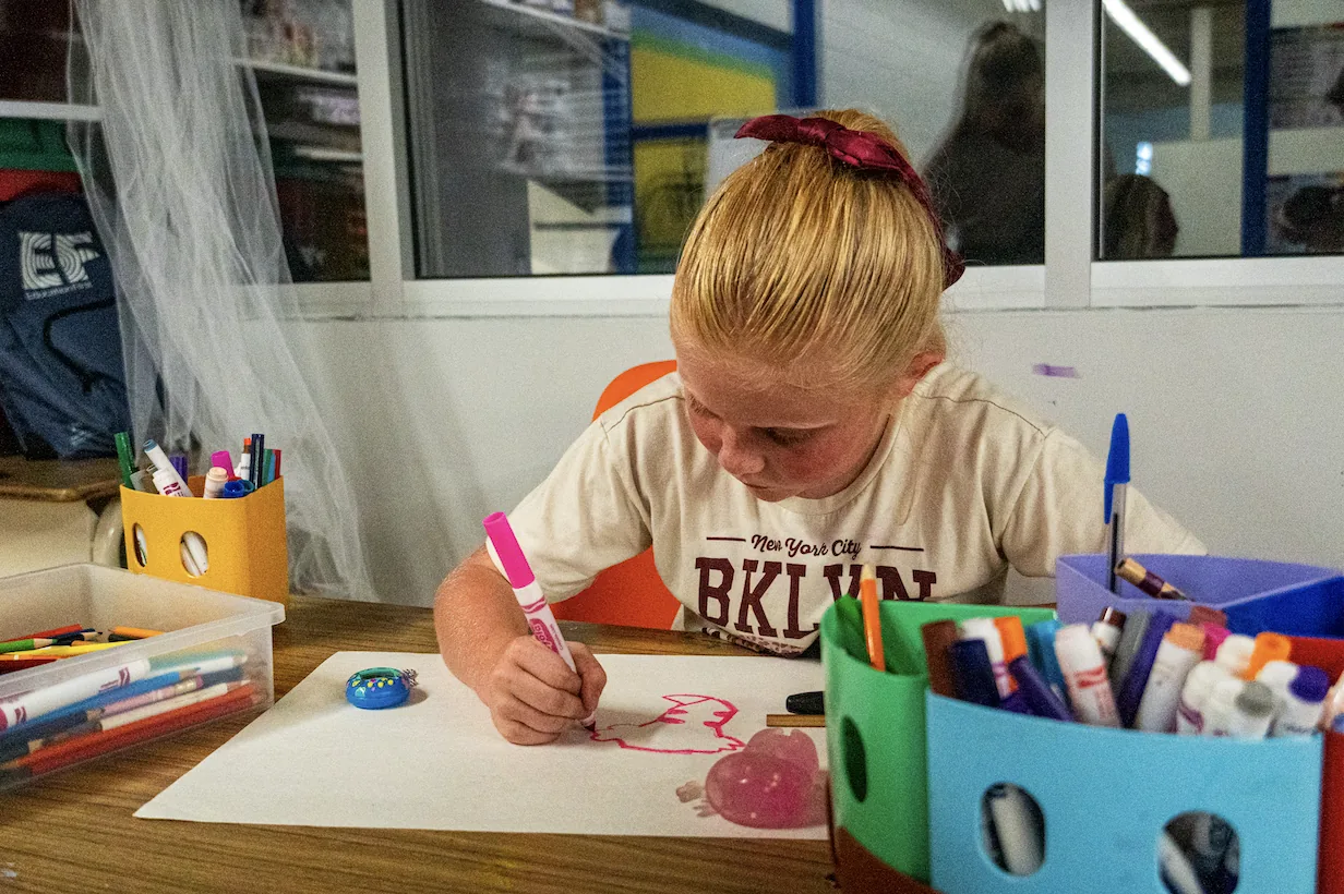 A girl drawing with markers at a craft table surrounded by art supplies