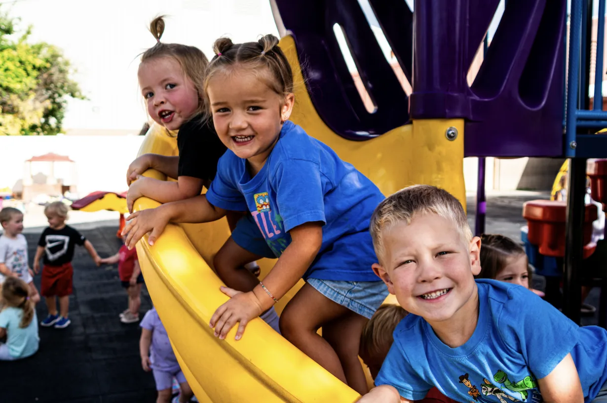 Three toddlers smiling together on playground equipment in the Kids Korner outdoor play area