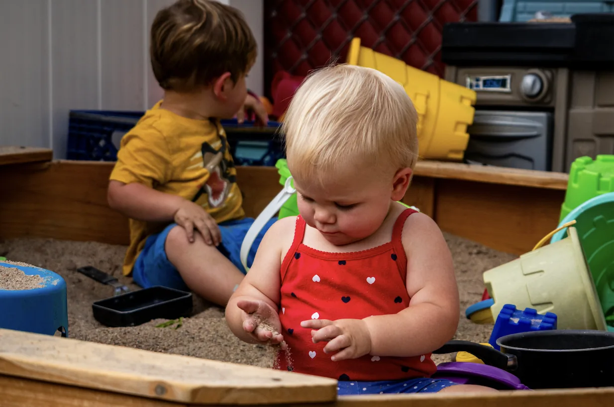 Toddlers playing together in an outdoor sandbox with buckets and toys at The Center