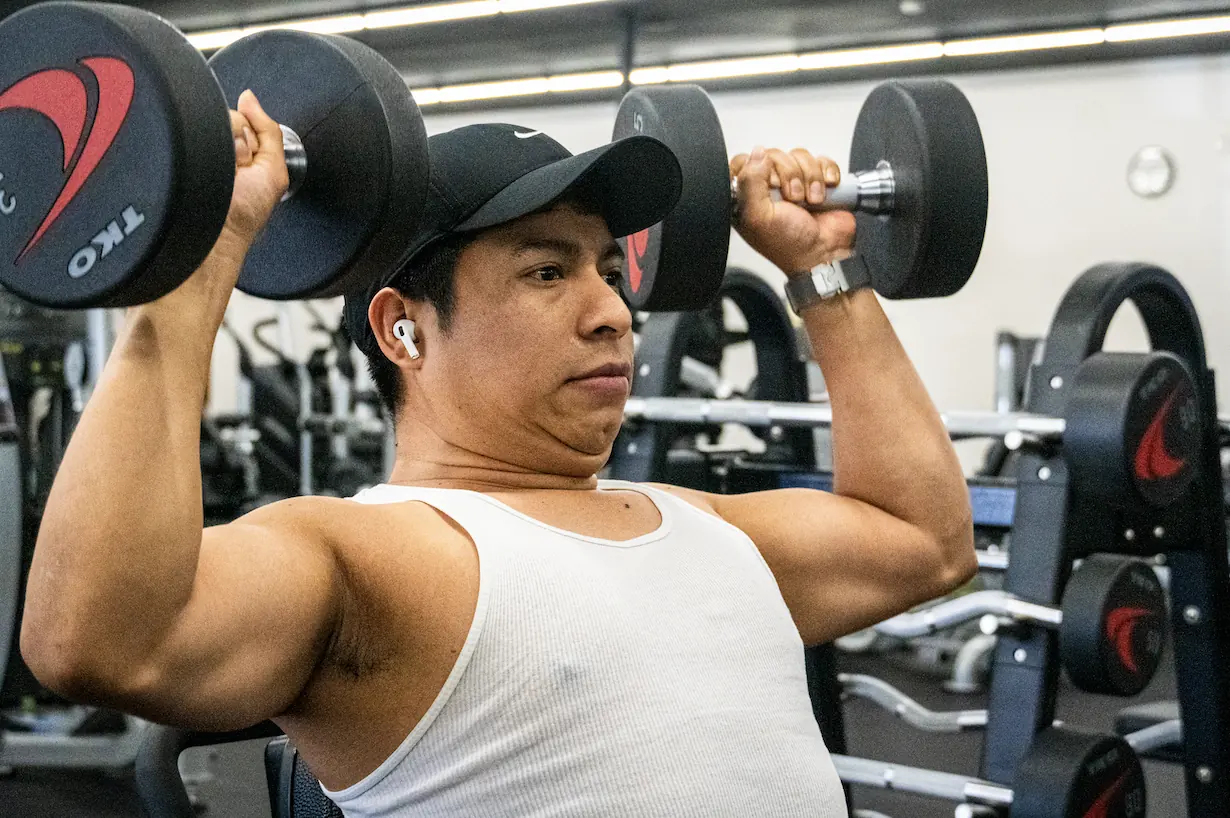 Member pressing dumbbells overhead beside the dumbbell racks at The Training Room